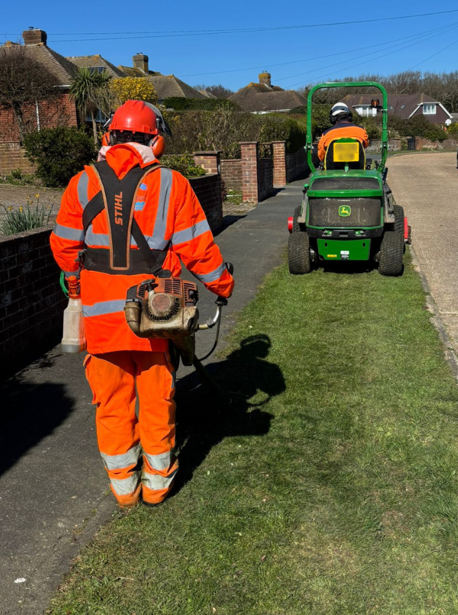 A man in high visibility clothing cutting a grass verge with a strimmer, following a man on a ride-on tractor style mower